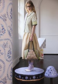 Kobieta w okularach, ubrana w kwiecistą koszulę, beżową spódnicę, fioletowe skarpetki i białe szpilki, trzymająca kwiecistą torbę na ramię, stojąca na ozdobionej fioletowej platformie wewnątrz budynku.