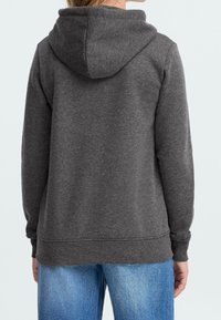 Personne portant un sweat à capuche gris foncé et un jean bleu, vue de dos sur un fond clair uni.