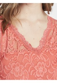 Korálový krajkový top s výstřihem do V, zdobený květinovými vzory a vlnitým okrajem. Měkká textura s detaily v několika vrstvách.