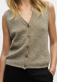 Bez piedurknēm, beigtas krāsas adīts žakets ar pogām, uzvilkts pār melnā svītrojuma biksēm, labajā rokā ir sudraba rokassprādze.