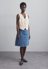 Beige linnen vest met knopen aan de voorkant, gecombineerd met een blauwe denim rok met een voorknipt en een rafelige zoom, gedragen met donkere instappers.