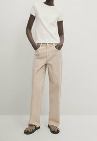 Beige levele ke culotte yhdistetty valkoiseen lyhythihaiseen t-paitaan. Asukokonaisuutta täydentävät ruskeat remmisandaalit. Yksinkertainen, minimalistinen muotoilu.