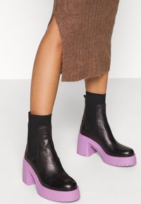 Ben i brun ribbad kjol med sidofickor, iförd svarta ankelstövlar med tjocka lila plattformshälar och elastiska sidopaneler.