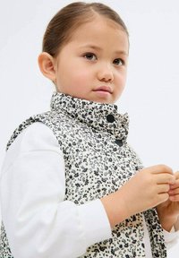 Hnedá vesta s kvetinovým vzorom v čierno-bielej farbe, s vysokým golierom, skombinovaná s bielou košeľou s dlhým rukávom. Obsahuje čierne gombíky a jemnú textúru.