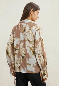 Lilleline pluus, mille värvivalikus on pruun ja valge, laiad varrukad, kitsendatud randmed ning sile ja kerge kangas.