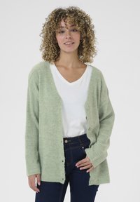 Cardigan en tricot vert clair avec une coupe ample, texture côtelée, encolure ronde et boutons sur le devant, porté sur un t-shirt blanc et un jean sombre.