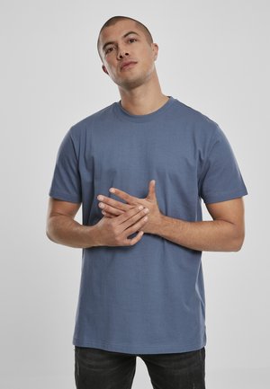 T-shirt di cotone blu con collo rotondo e maniche corte, caratterizzato da una vestibilità comoda. Indossato con jeans scuri, mani incrociate davanti.
