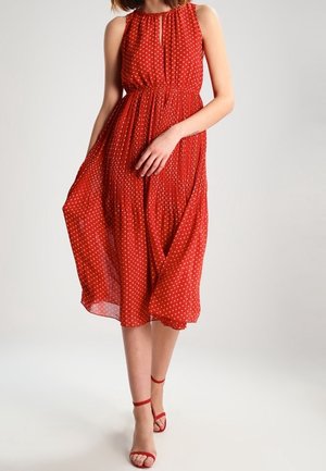 Femme portant une robe midi rouge sans manches à pois blancs et des sandales rouges à talons hauts avec bride à la cheville, debout devant un fond uni.