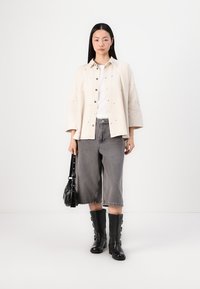 Giacca camicia di colore crema chiaro con chiusura a bottoni, abbinata a pantaloni denim grigi a gamba larga e stivali neri sopra il ginocchio. Inclusa una borsa nera.