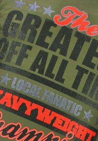 Groen stof met tekst in zwart, rood en grijs die leest "The Greatest of All Time," "Local Fanatic," en "Heavyweight" met sterren erboven en eronder.