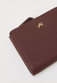 Portefeuille en cuir bordeaux avec une finition texturée, arborant un accent logo en métal doré. Design compact avec une fermeture éclair.