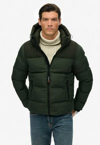 Mørkegrøn pufferjakke med lynlås, hætte og ribbet krave; har vandrette baffler og to frontlommers med lynlås. Bæres over en lys sweater.