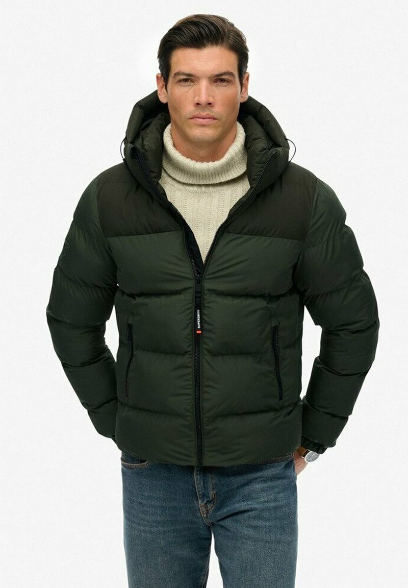 Mørkegrøn pufferjakke med lynlås, hætte og ribbet krave; har vandrette baffler og to frontlommers med lynlås. Bæres over en lys sweater.