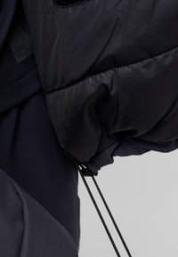Veste rembourrée en marine foncé avec une texture brillante, dotée de poignets élastiques, d'un cordon de serrage à l'ourlet et d'une étiquette de logo visible sur le tissu.