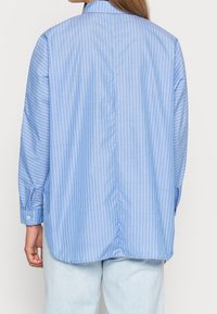 Chemise à rayures bleues à boutons avec un empiècement au dos et des manches longues, présentant des rayures blanches verticales et des boutons aux poignets. Coupe ajustée avec un ourlet arrondi.