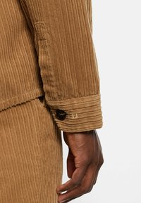 Braune Cordjacke mit vertikaler Rippung, einem knöpfbaren Ärmel und einer weichen Textur, mit sichtbaren Steppdetails.