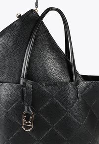 Zwei schwarze, strukturierte Taschen sind übereinander gestapelt; eine größere Tote mit langen Griffen und eine kleinere, gefaltete Tasche darin, die ein Logodetail aufweist.