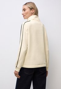 Donna con capelli biondi corti che indossa un maglione crema a collo alto con strisce nere sulle maniche e jeans blu scuro, voltata di lato.