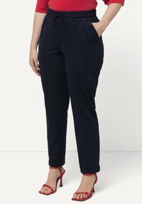 Pantalones azul marino de cintura elástica con bolsillos laterales y un bolsillo de cargo; fabricados en tela suave; diseño ajustado; combinados con tacones rojos de tiras.