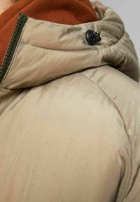 Beżowa kurtka puchowa o gładkiej fakturze, z zielonym zamkiem błyskawicznym i czarnym zaczepem na kapturek, nałożona na pomarańczowy element odzieży.