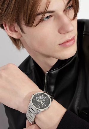 Orologio in acciaio inossidabile argento con quadrante nero, tre sotto-quadranti e bracciale a maglia testurizzata, indossato su un polso contro una giacca nera.