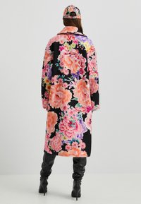 Dlouhý květinový kabát v černé barvě s velkými růžovými a fialovými květy, s výrazným límcem a měkkou texturou. Doplněn o ladící čepici.