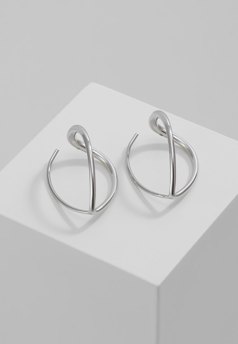 Paire de boucles d'oreilles argentées torsadées et ouvertes, présentées sur un piédestal blanc avec un fond gris minimaliste.