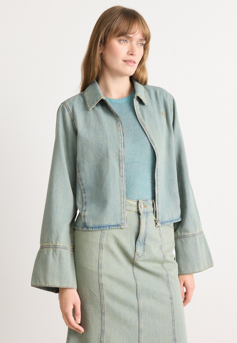 Lyseblå denimjakke med glidelås foran, spiss krage og klokkeslett, kombinert med en matchende lyseblå skjørt og teksturert topp.