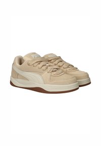 Beige suède sneakers met dikke veters, witte zijstrepen, gevoerde kraag en een bruine rubberen zool, zij aan zij geplaatst op een witte achtergrond.