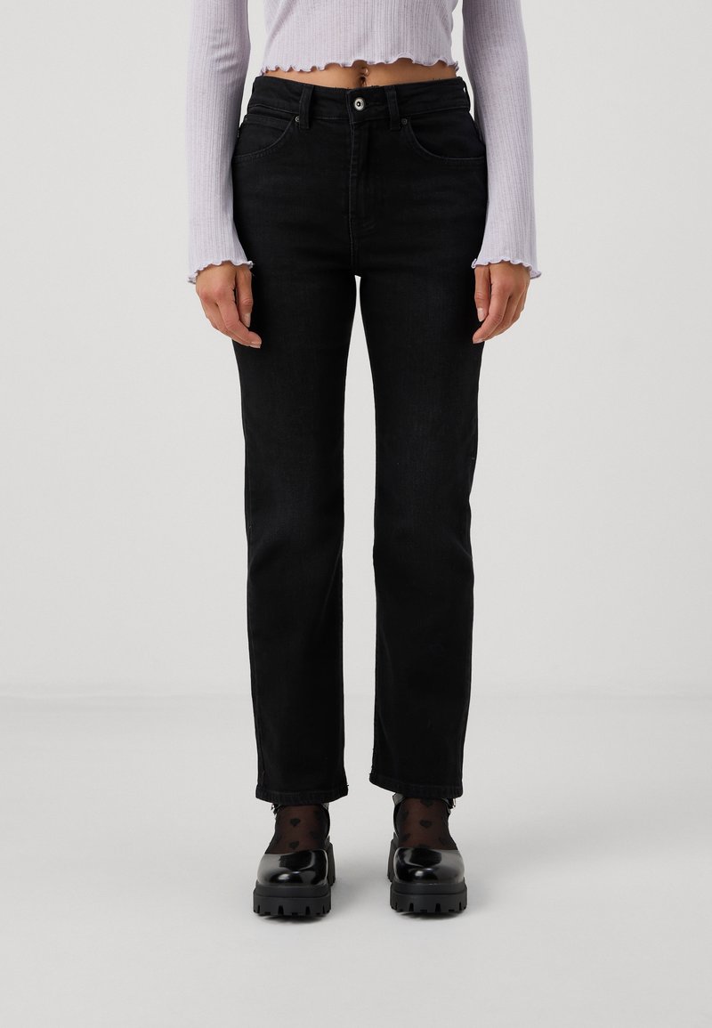 Personne portant un jean noir droit taille haute, des chaussettes noires transparentes avec motif cœur, des chaussures noires épaisses et un top court blanc à manches longues.