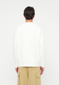 Mężczyzna w prostym białym długim swetrze i beżowych spodniach cargo, stojący tyłem na białym tle.