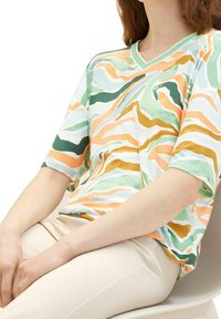 Femme assise portant un t-shirt ample à col en V avec des rayures abstraites vertes, orange et blanches et un pantalon beige.