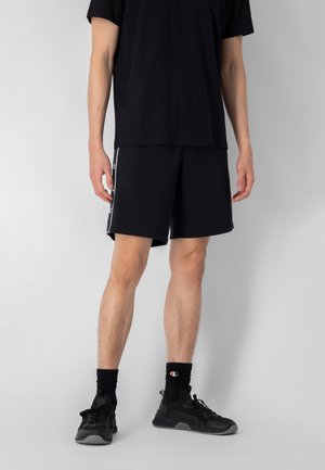 Pantalones cortos negros con rayas laterales blancas, combinados con una camiseta negra. Modelo lleva zapatillas deportivas negras y calcetines negros. Fondo gris liso.