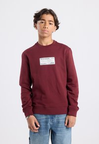 Burgundy sweatshirt with a round neckline, ribbed cuffs, and a front label patch featuring text. Worn with light blue denim pants.