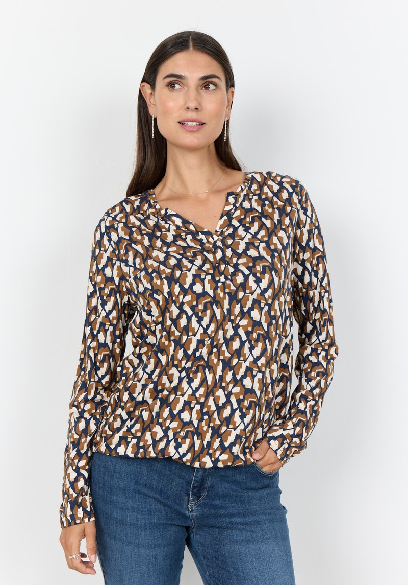 Langärmlige Bluse mit einem geometrischen Muster in Braun-, Weiß- und Blautönen, mit V-Ausschnitt und weichem Stoff. Kombiniert mit blauen Jeans.