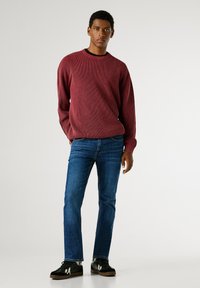Bordový pletený svetr s kulatým výstřihem, kombinovaný s modrými džíny a černými teniskami. Svetr má žebrovanou texturu a manžety.