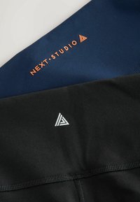 Marineblå og sort træningstøj med orange "NEXT.STUDIO" logo og et hvidt geometrisk emblem. Glat tekstur, tætsiddende design og kontrasterende farver.