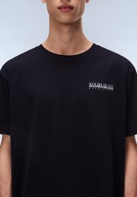Black cotton t-shirt with short sleeves, featuring a small white logo on the chest. Round neckline and soft texture, minimalist design.