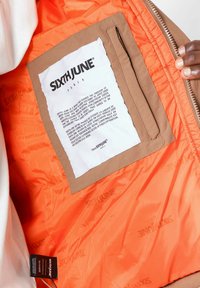 Voering van gewatteerde jas in fel oranje met een bruine stoffen zak. Witte label met zwarte tekst met het merk en onderhoudsinstructies.