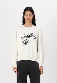 Felpa bianca con girocollo e testo ricamato nero "Saddle Up", con spalle scese e polsini a costine, abbinata a pantaloni grigio scuro.