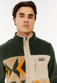 Mann in einer grün-beige Fleecejacke mit geometrischen Mustern und einer mit "PRO TEST" beschrifteten Reißverschluss-Brusttasche.