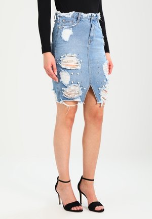 Femme portant une jupe en jean bleu usé avec des ornements en perles, une fente à l'avant, associée à des sandales noires à talons hauts et un top noir à manches longues.