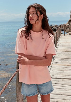 Kvinde med langt mørkt hår iført en pink skjorte og denimshorts står på en træbro ved havet, armene krydsede, kigger mod venstre.