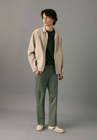 Light beige jacket with a relaxed fit, black t-shirt underneath, slim green trousers, and cream sneakers with green accents.