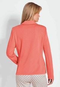 Koraalblazer met een klassieke kraag, getailleerde pasvorm, lange mouwen en structuurstof, gecombineerd met met patroondachtige broek met druppelontwerpen.