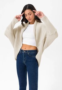 Beige, gerippter Strickcardigan mit offener Vorderseite, überschnittenen Schultern und weiten Ärmeln. Getragen über einem weißen Crop-Top und dunklen Jeans.