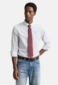 Vit randig skjorta med knappar, rött slips med paisleymönster, blå jeans, brunt bälte, stående pose, händerna på höfterna.