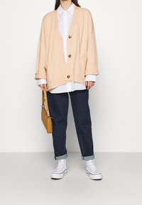 Meruňkový pletený kardigan s výstřihem do V, volným střihem, třemi knoflíky a předními kapsami, doplněný bílou košilí a tmavými džínami.