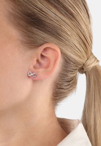 Orecchino a perno argento a forma di infinito con piccoli cristalli incastonati, indossato sull'orecchio sinistro di una persona con capelli biondi raccolti.