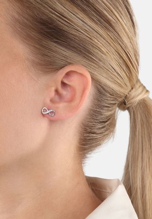 Silberner Ohrstecker in Form einer Unendlichkeit mit kleinen eingelassenen Steinen, getragen am linken Ohr einer Person mit zurückgegebundenem blonden Haar.